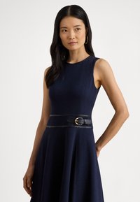 Marineblå ermeløs kjole med vidt skjørt, som har et kontrastfarget svart belte med gullspenne i midjen. Glatt tekstur, tettsittende design.