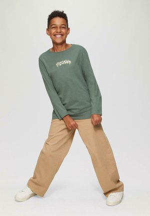 Langärmliges grünes Baumwoll-T-Shirt mit "FREEMIND" Grafik, beigen baggy Hosen und weißen Sneakers. Kind posiert spielerisch.