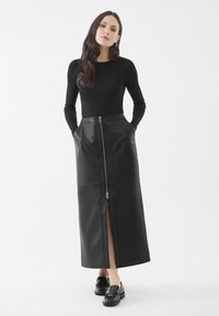 Vrouw staat met handen in de zakken, gekleed in een zwart langemouwen top, zwarte leren midi rok met rits aan de voorkant, en zwarte instapschoenen, op een witte achtergrond.