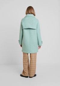 Cappotto in lana verde menta con un ampio colletto e design a pattina; abbinato a pantaloni a quadri marroni, che mostrano una vestibilità comoda e una consistenza morbida.