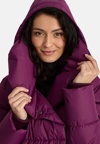 Violeta puffy jaka ar pārmērīgu apkakli, pogu detaļām un ribotām piedurknēm. Gluda, spīdoša auduma ar pildījuma dizainu siltumam.