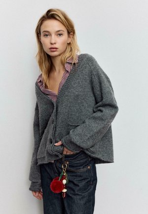 Ylimitoitettu harmaa villatakki, jossa syvä V-kaula-aukko, resorit hihansuissa ja etutaskut, kerrospukeutunut raidallisen paidan päälle. Mukana avaimenperä.