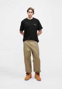 Camiseta de algodón negra con logo en el frente, pantalones khaki de pierna ancha y botas marrones con cordones. El modelo lleva una gorra beige y está de pie con las manos detrás de la espalda.