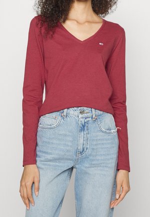 Vrouw draagt een nauwsluitend langarmig bordeauxrood V-hals topje, ingestopt in een lichtblauwe, hooggetailleerde spijkerbroek met zichtbare knopen en zakken.