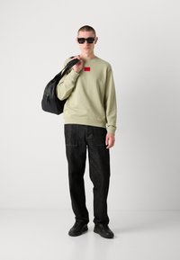 Sweatshirt vert clair avec un patch logo rouge, associé à un pantalon en denim foncé et des baskets noires. Le modèle porte un sac noir sur une épaule.