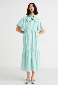Vestido verde menta de largo a la rodilla con mangas acampanadas, cuello con volantes y un lazo en el cuello. Tejido suave con cintura fruncida y falda en capas.