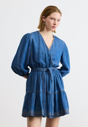 Tânără purtând o rochie tip wrap din denim albastru, cu mâneci bufante și o centură legată în talie, stând pe un fundal alb simplu.