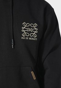 Mustat hupparit, jotka on valmistettu puuvillasta ja joissa on etutasku. Hupparissa on kultalangalla brodattu logo "PAS DE MONACO" ja monimutkainen kuvio rinnassa.