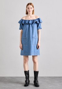 Svijetloplava denimska haljina s otkopčanim ramenima, volanima oko vrata, dva prednja džepa, ravnog kroja, uparena s crnim čizmama do gležnja.