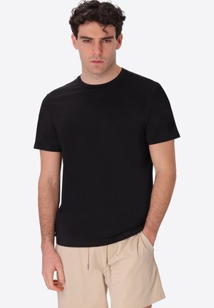 Uomo con capelli scuri e ricci che indossa una maglietta nera semplice a maniche corte e pantaloncini beige, posando con una mano in tasca su sfondo bianco.