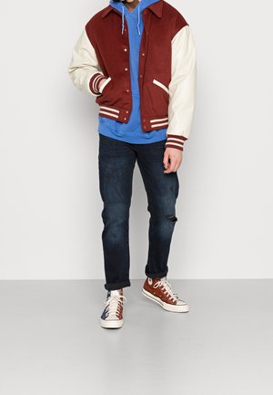 Chaqueta universitaria roja y crema con puños acanalados, llevada sobre una sudadera con capucha azul. Combinada con vaqueros oscuros y zapatillas marrones, blancas y azules.