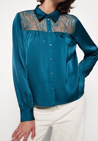 Teal satinblus med en genomskinlig spetspanel över bröstet, långa puffärmar och knäppning. Slät textur med en skräddarsydd passform.