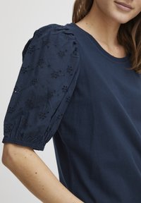 T-shirt bleu marine avec des manches bouffantes, ornée de broderies florales sur le tissu des manches, col rond, et texture en coton doux.