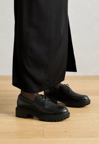 Zwarte leren schoenen met een ronde neus, dikke rubberen zool en veters. Het ontwerp heeft decoratieve stiksels langs de bovenkant.