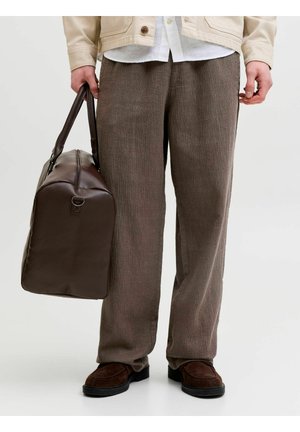 Personne portant un pantalon marron texturé, des chaussures marron foncé, une veste beige, tenant un grand sac de voyage en cuir marron foncé à ses côtés.