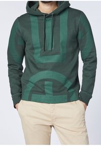 Felpa con cappuccio di un verde scuro, decorata con un grande motivo geometrico di un verde più chiaro. Dotata di cordini e polsini a costine. Tessuto di finitura liscia.