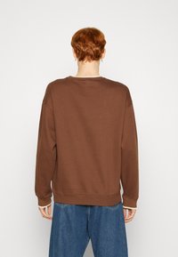 Hnědá mikina s texturovanou tkaninou, volným střihem, žebrovanými manžetami a spodním lemem. Má zaoblený výstřih a jemné kontrastní stehy.