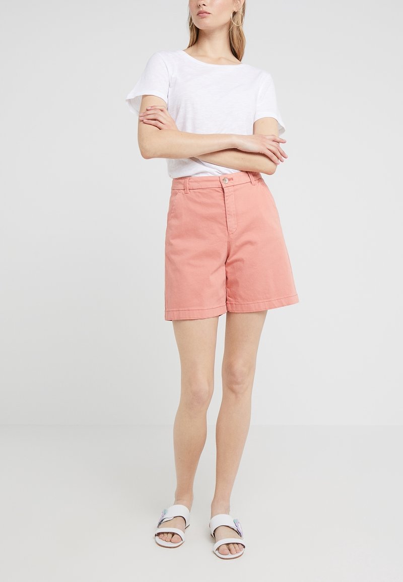 Femme debout, les bras croisés, portant un haut blanc à manches courtes, un short rose taille haute et des sandales blanches avec des détails floraux.