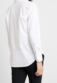 Weiße Button-up-Hemd mit langen Ärmeln, yoke Naht hinten, abgerundetem Saum und Knopfmanschetten; aus glattem Stoff mit matter Textur.