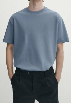 Person trägt ein schlichtes blaues Kurzarm-T-Shirt, das in hoch taillierte schwarze Hosen gesteckt ist, die Hände in den Taschen, von Hals bis Taille gezeigt.