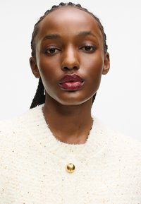 Balts teksturēts žakets ar apaļu kakla izgriezumu, rotāts ar zeltainām pērlītēm un zelta pogu akcents. Gluda auduma ar maigu izskatu.