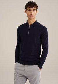 Polo de punto azul marino con cuello de cremallera, mangas largas y puños acanalados. Combinado con pantalones gris claro, que presentan un diseño casual.