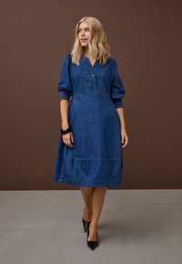 Blond ínka v modrom rifľovom midi šatách a čiernych lodičkách stojaca pri hnedej stene a sivom podlahe.