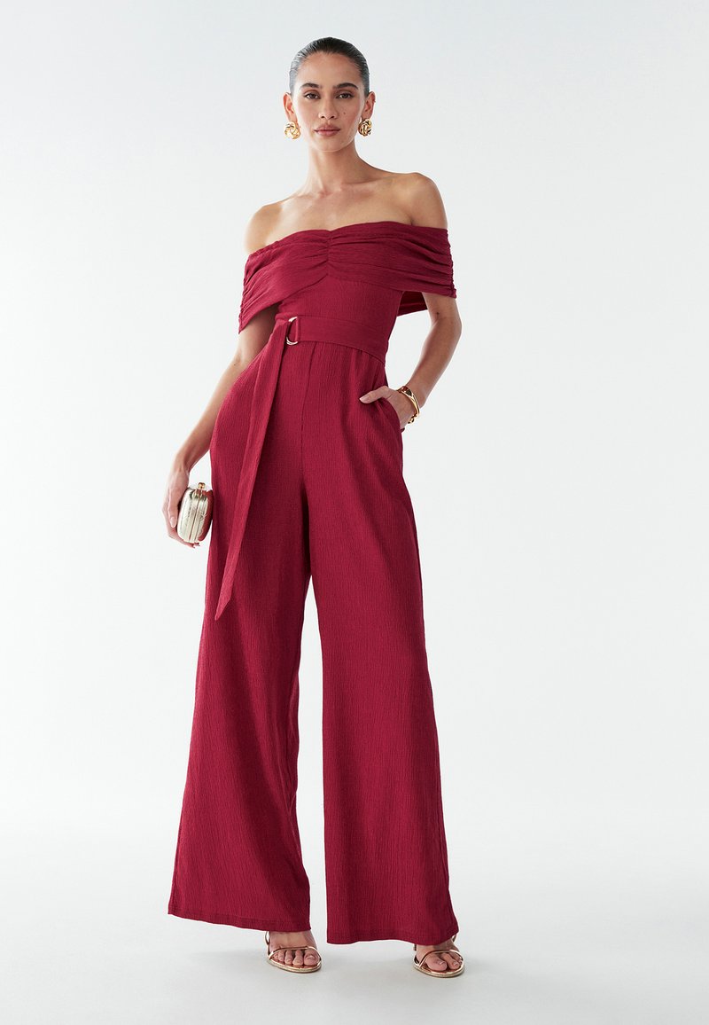 Červený overal s odhalenými ramenami z textúrovanej látky, so širokými nohavicami, opaskom v páse a jemným leskom, doplnený o sandále s remienkami.