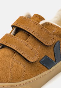 Zapatillas de ante marrón con dos correas de velcro, una superficie texturada y un acento de goma negra en el costado. Forro suave visible en el interior.