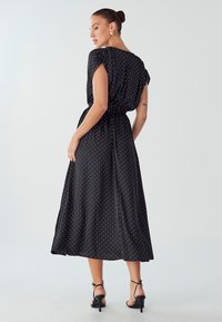 Black polka dot dress with short sleeves, cinched waist, and flowing skirt. Silky texture and mid-calf length, paired with strappy black heels.