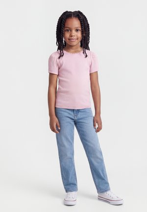 Νεαρή κοπέλα με πλεξούδες, φορώντας ροζ κοντομάνικη μπλούζα, ανοιχτό μπλε τζιν και λευκά αθλητικά, στέκεται μπροστά σε λευκό φόντο.