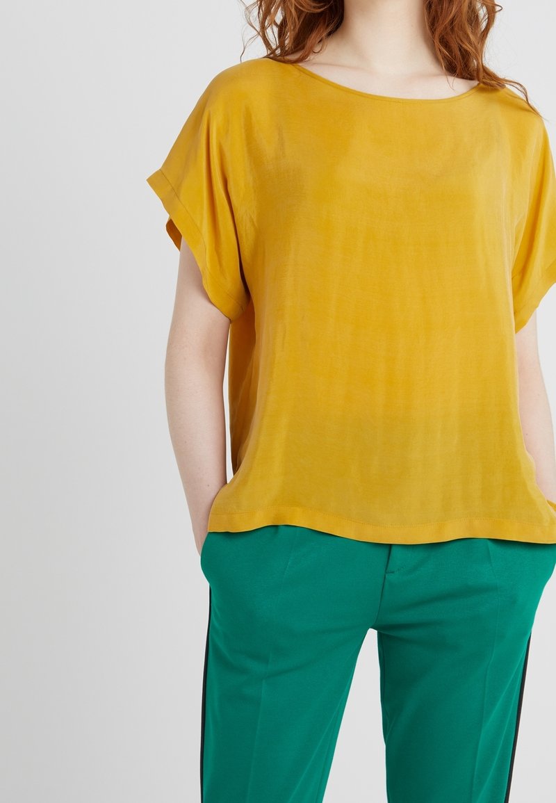 Top a maniche corte color giallo senape in tessuto leggero con vestibilità ampia. Abbinato a pantaloni verdi con strisce laterali nere.