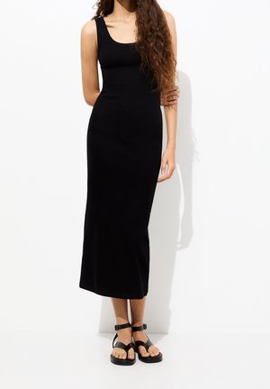 Kobieta w czarnej dopasowanej sukience midi bez rękawów i czarnych płaskich sandałach stoi przy białej ścianie.
