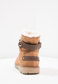 Bottine en daim marron avec doublure en fausse fourrure blanche, sangle en cuir marron, accent métallique rond et semelle en caoutchouc texturée.