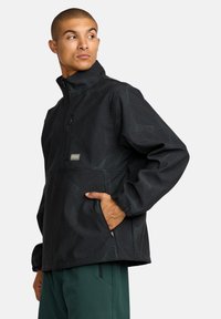 Schwarze Pulloverjacke mit kurzem Reißverschluss und strukturiertem Wellenmuster, ausgestattet mit einer Fronttasche, elastischen Bündchen und einem lockeren Schnitt, kombiniert mit grünen Hosen.