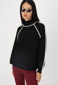Pull noir avec des détails blancs, col montant et manches bouffantes. Associé à un pantalon fitted bordeaux. Texture lisse.