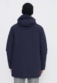 Giacca imbottita blu navy con cappuccio, caratterizzata da un design trapuntato, maniche lunghe e lunghezza media. Il materiale sembra liscio e resistente all'acqua.