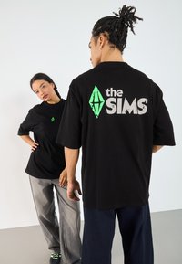 Sorte oversized t-skjorter med et grønt diamantsymbol og "the SIMS" i grått, med en glatt tekstur og korte ermer.
