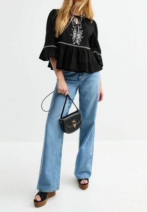 Vrouw draagt een zwarte blouse met bloemenborduursel, lichtblauwe wijde jeans, zwarte plateauzandalen, en houdt een kleine zwarte handtas vast.