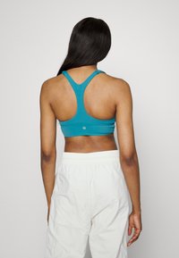 Sutiã desportivo teal com um design às costas em raçador, textura suave e um pequeno detalhe de logo na parte traseira, combinado com calças brancas de cintura elástica.