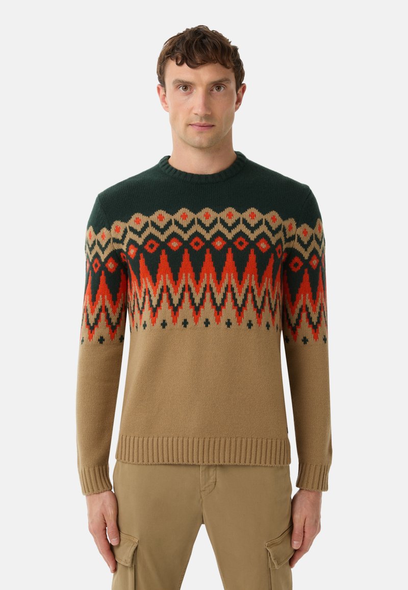 Man wearing tan cargo pants and a crewneck sweater with green, red, black, and tan geometric patterns across the chest and sleeves.