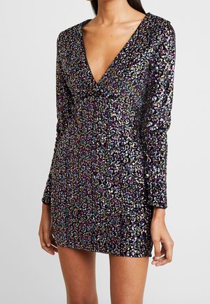 Vrouw die een mini-jurkje met lange mouwen en een diepe V-hals draagt, bedekt met meerkleurige pailletten op een zwarte achtergrond.