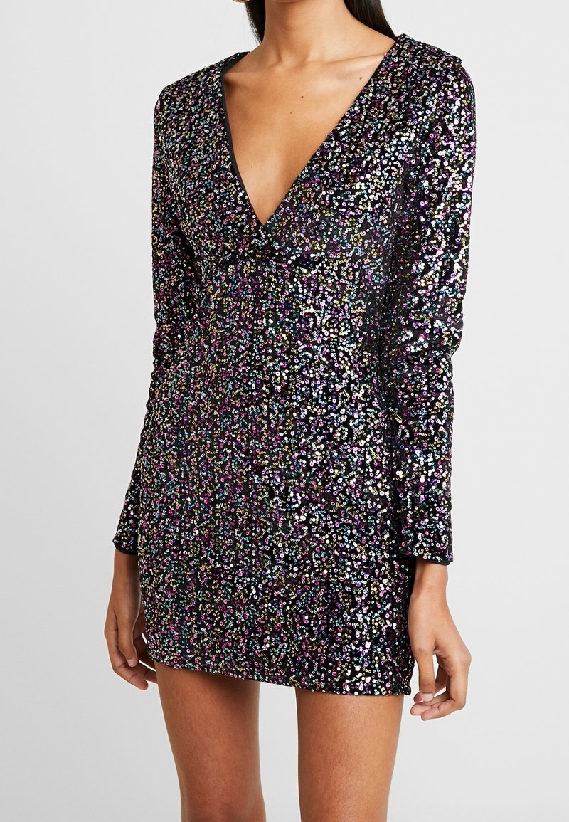 Femme portant une mini-robe à manches longues avec un décolleté en V profond, ornée de sequins multicolores sur un fond noir.