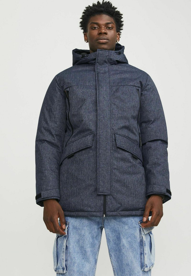 Chaqueta acolchada de color azul marino con un alto cuello y capucha, que cuenta con dos bolsillos frontales y cierre de cremallera, hecha de un tejido texturizado y resistente al agua.