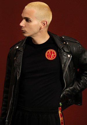 Zwarte t-shirt met rood cirkelvormig logo, gedragen onder een gestructureerde zwarte leren jas. Het model heeft kort blond haar en kijkt zijwaarts.