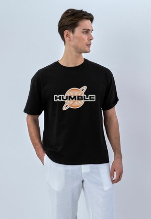 Mann trägt schwarzes T-Shirt mit orangefarbenem Globus und dem Wort „HUMBLE“ bedruckt, kombiniert mit weißer Hose, schaut nach rechts vor einfachem Hintergrund.