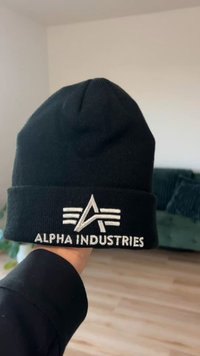 Gorro de malha preto com bainha dobrada. Apresenta um logótipo bordado a prata com letras estilizadas e detalhes a dizer "ALPHA INDUSTRIES".
