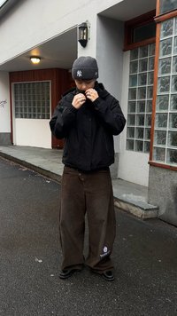 Sort lynlåstrøje, grå strikket hue med logo, brune vidde ben fløjl bukser, skinnende sorte sko. Urban udendørs miljø med beton og glas.