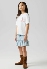 T-shirt en coton blanc avec un graphique "REINE DU DÉSERT", associé à une jupe en denim bleu clair avec un ourlet volé et des bottes en daim marron.