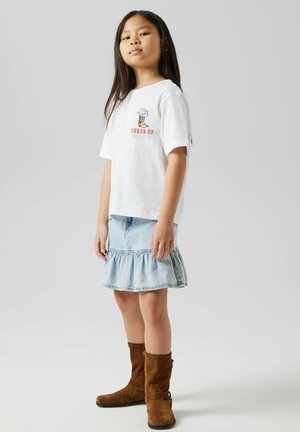 T-shirt en coton blanc avec un graphique "REINE DU DÉSERT", associé à une jupe en denim bleu clair avec un ourlet volé et des bottes en daim marron.